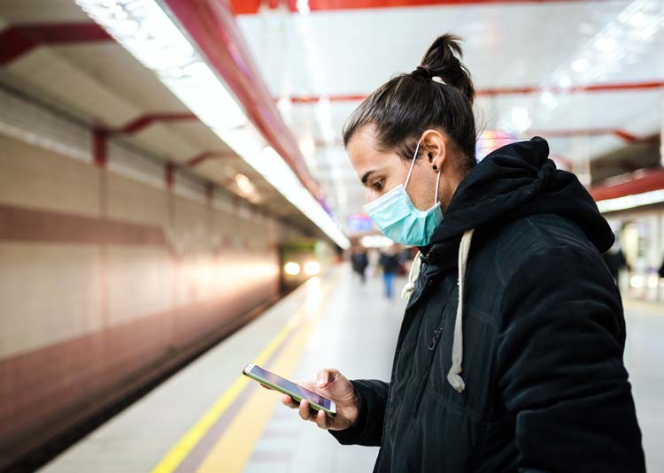 Mann mit Mundschutz steht an U-Bahn-Station und schaut auf Mobiltelefon